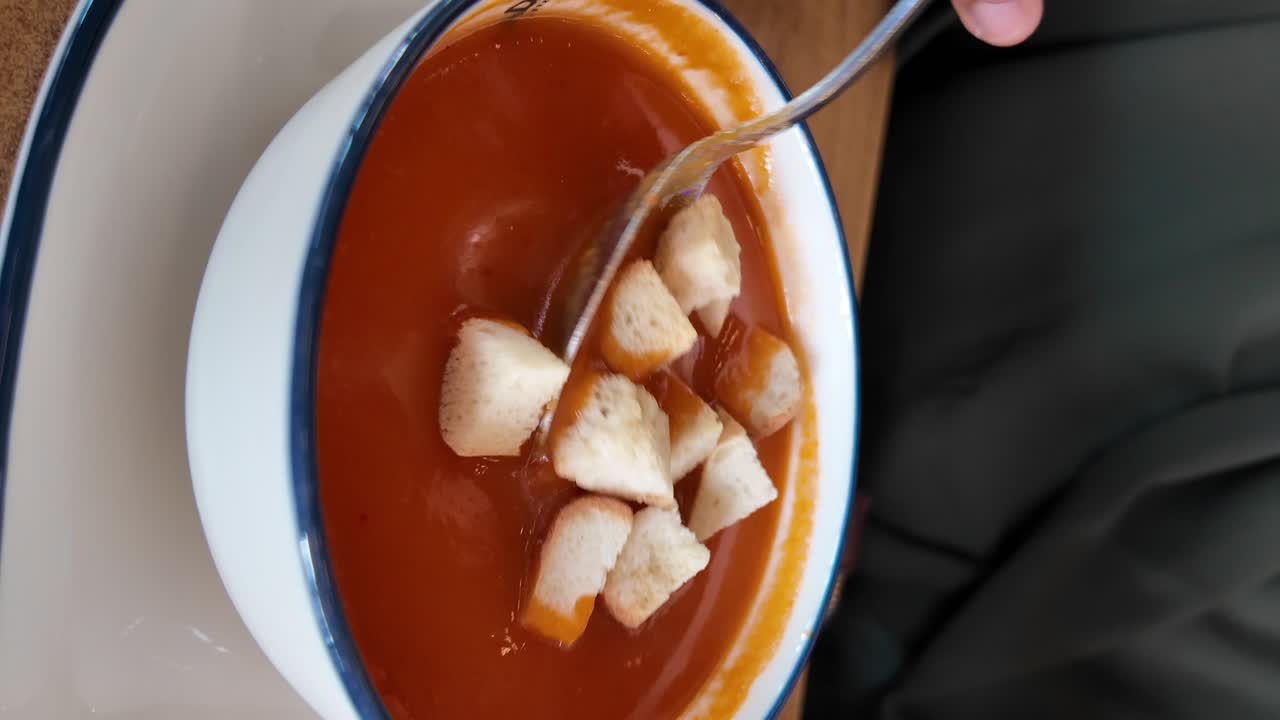 Close-up of tomato soup with croutons in a white bowl being stirred with a spoon