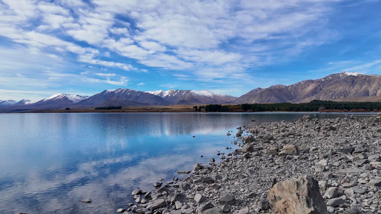 Drone footage captures Lake Tekapo's serene waters and rocky shoreline under a vibrant sky, framed by distant mountains