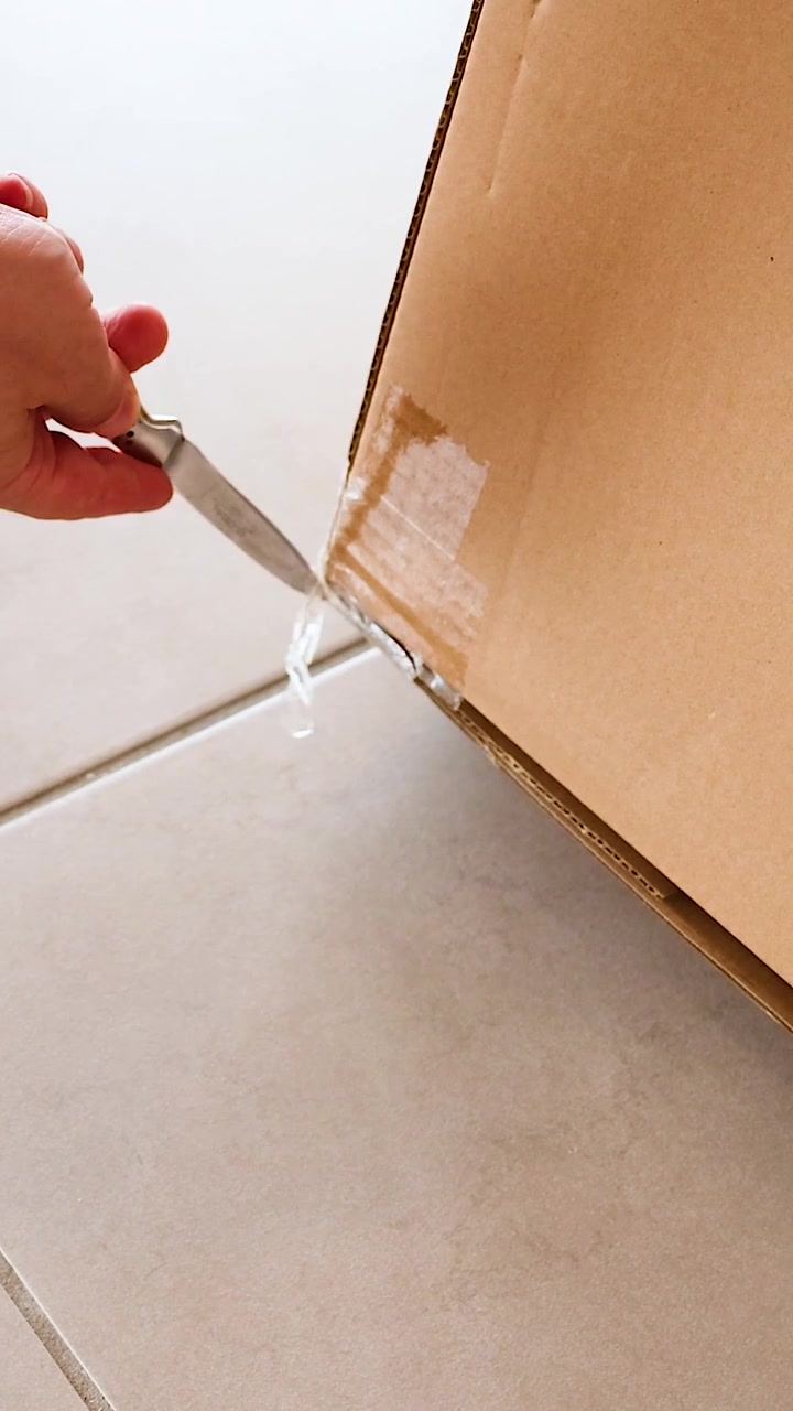 A person carefully cuts open a cardboard box using a knife on a tiled floor, in bright lighting