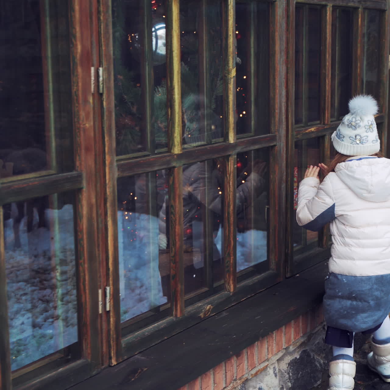 Little girl is looking through the window into the house outdoors. Small child in warm clothes standing near the windows in winter.