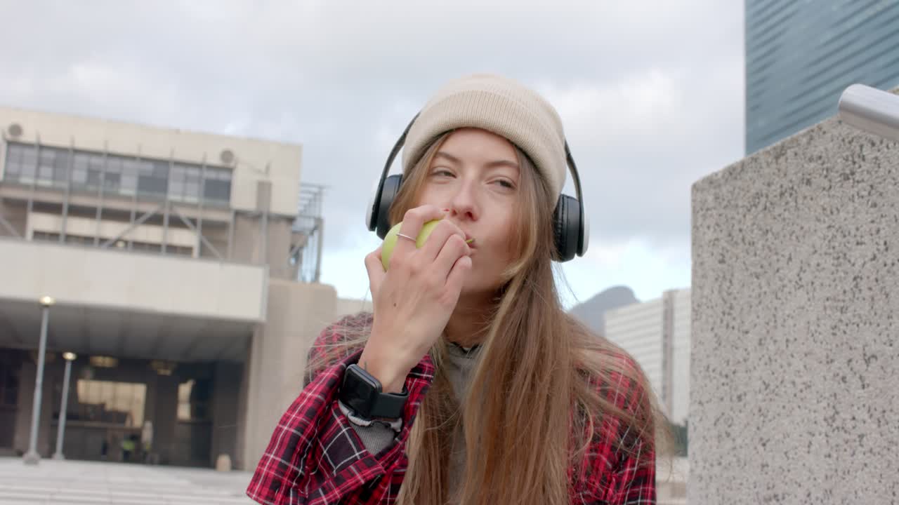Mujer con auriculares mordiendo una manzana verde en un saliente de hormigón en una plaza urbana, durante una pausa para el tentempié.