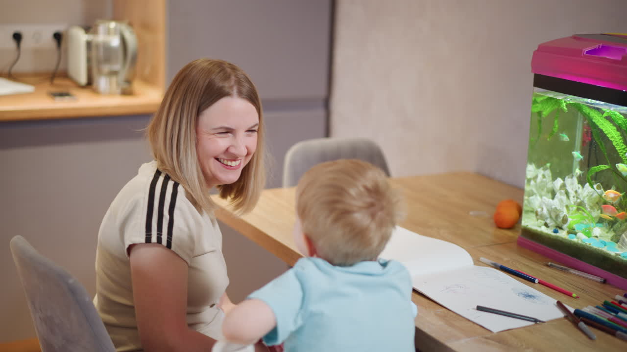 Happy woman smiling and playing with young child at home sitting near desk with notebooks, pens, aquarium, colorful fish, showing love, joy, bonding, laughter, affection, family connection