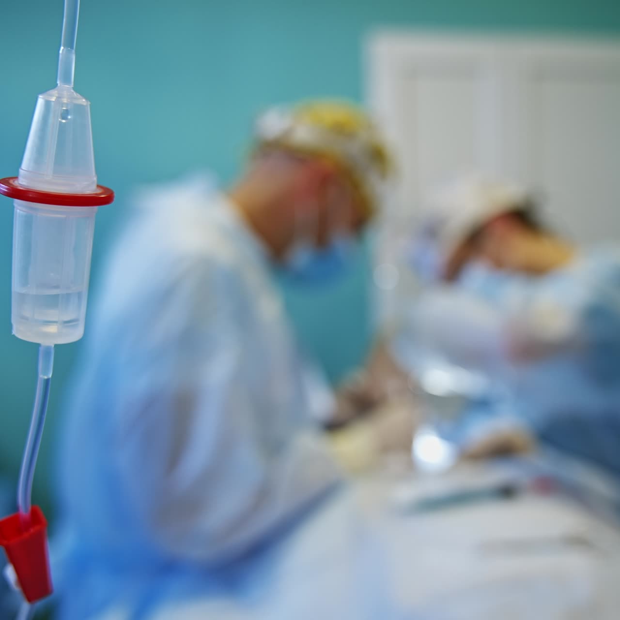 Drop counter with liquid dripping inside. Close up. A couple of surgeons performing surgery at backdrop in blur