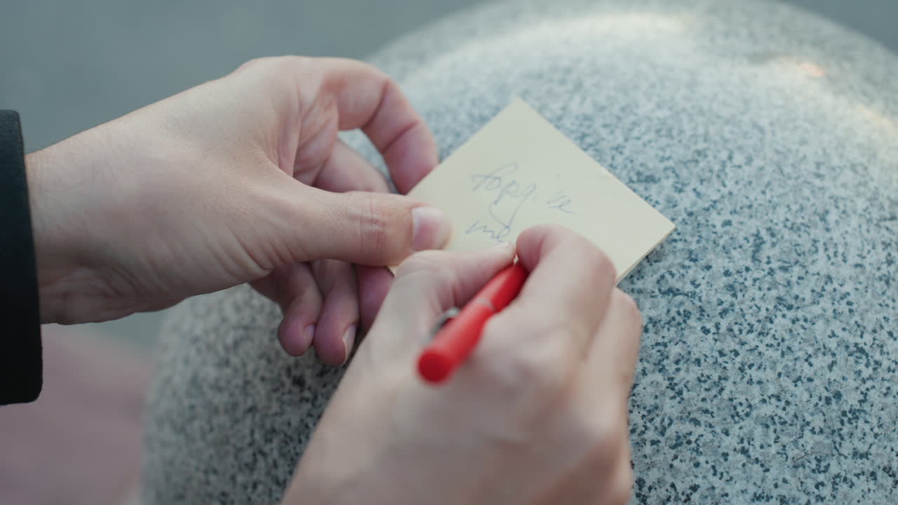 Closeup hands writing note on granite, red pen on sticky paper against rounded bollard coat sleeve visible, deliberate strokes, quick doodle and listmaking, planner leaving reminder, quiet urban