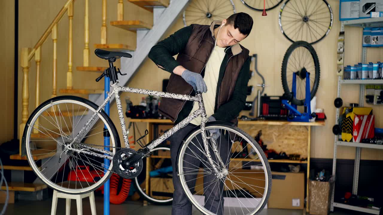 Young maintenance man is assembling bicycle placing stearing wheel and fixing it while listening to music with earphones in his workplace. Repair and people concept.
