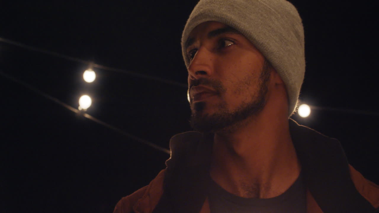 밤에 지붕 위에 있는 예쁜 젊은 히스패닉 남자의 초상화. 배경에 빛이 있는 심각한 모습을 하고 있다.