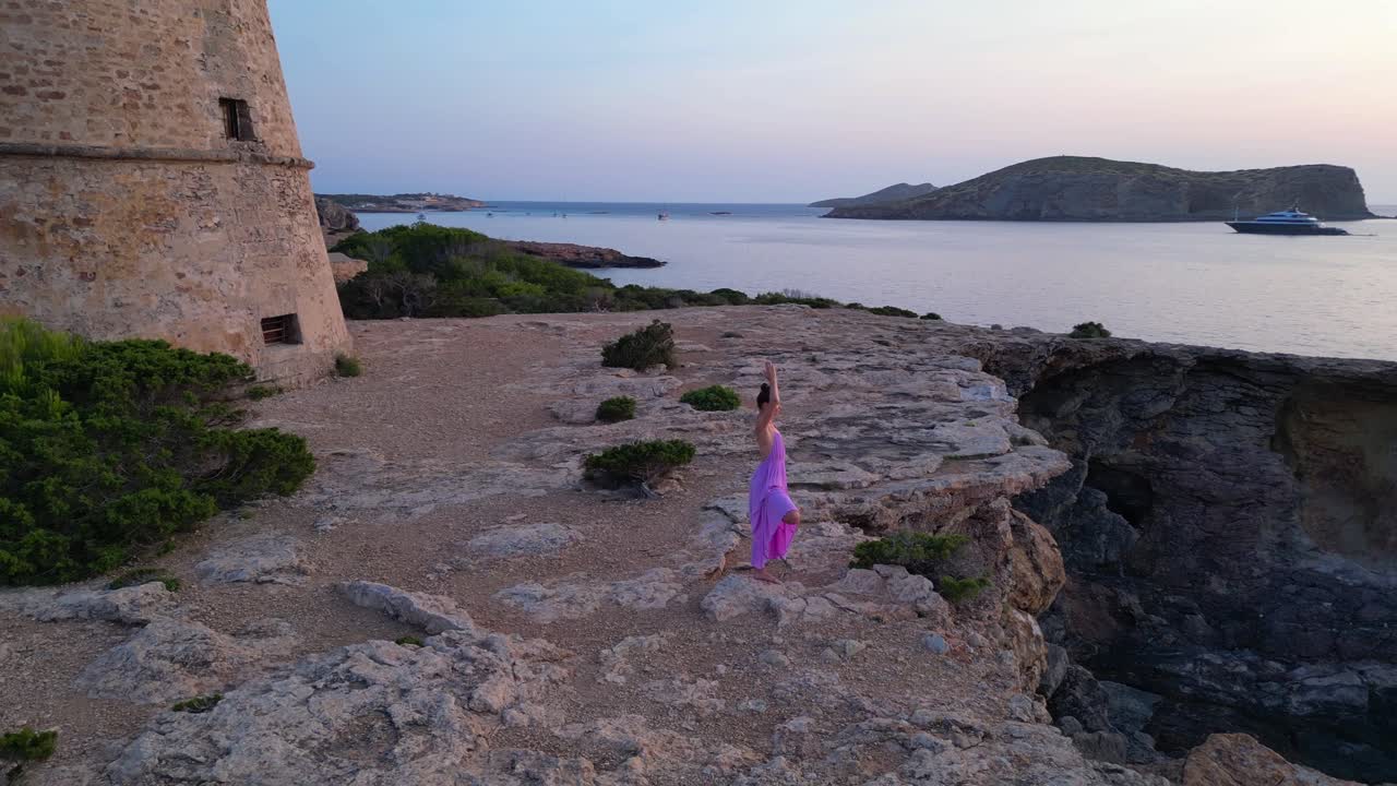 Woman in yoga pose enjoying the sunset near Torre d'en Rovira, a defense tower built in the 18th century in Cala Comte, Ibiza, Spain. Great aerial view flight panorama orbit drone