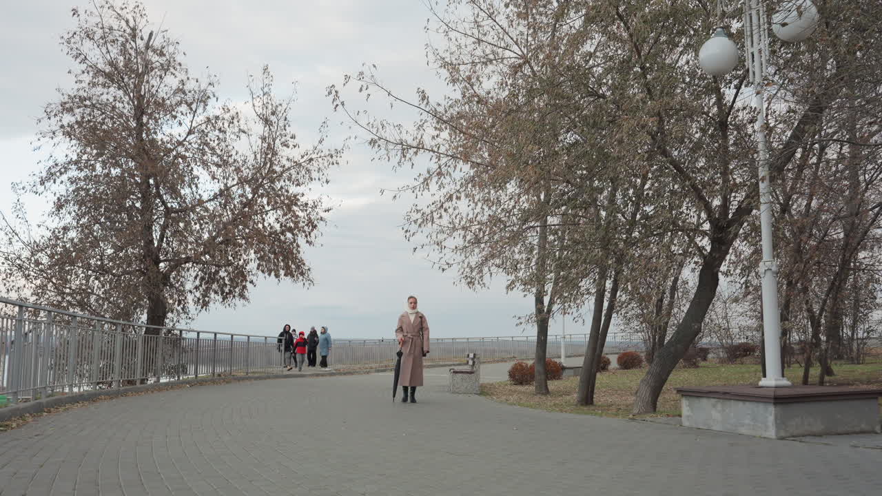 Femme caucasienne marchant vers un belvédère sous un ciel nuageux, avec un long manteau et une écharpe, un lampadaire et une balustrade encadrant la promenade, des touristes au loin regroupés près de la rambarde, ambiance de rencontre décontractée, scène urbaine prise sur le vif.