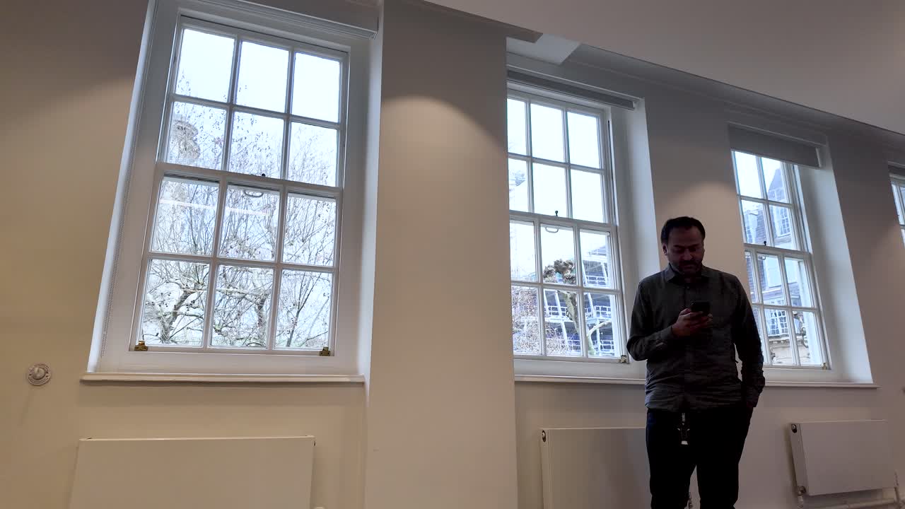 Un uomo che passeggia accanto ad alte finestre d'ufficio, controllando il suo smartphone durante una breve pausa di lavoro.