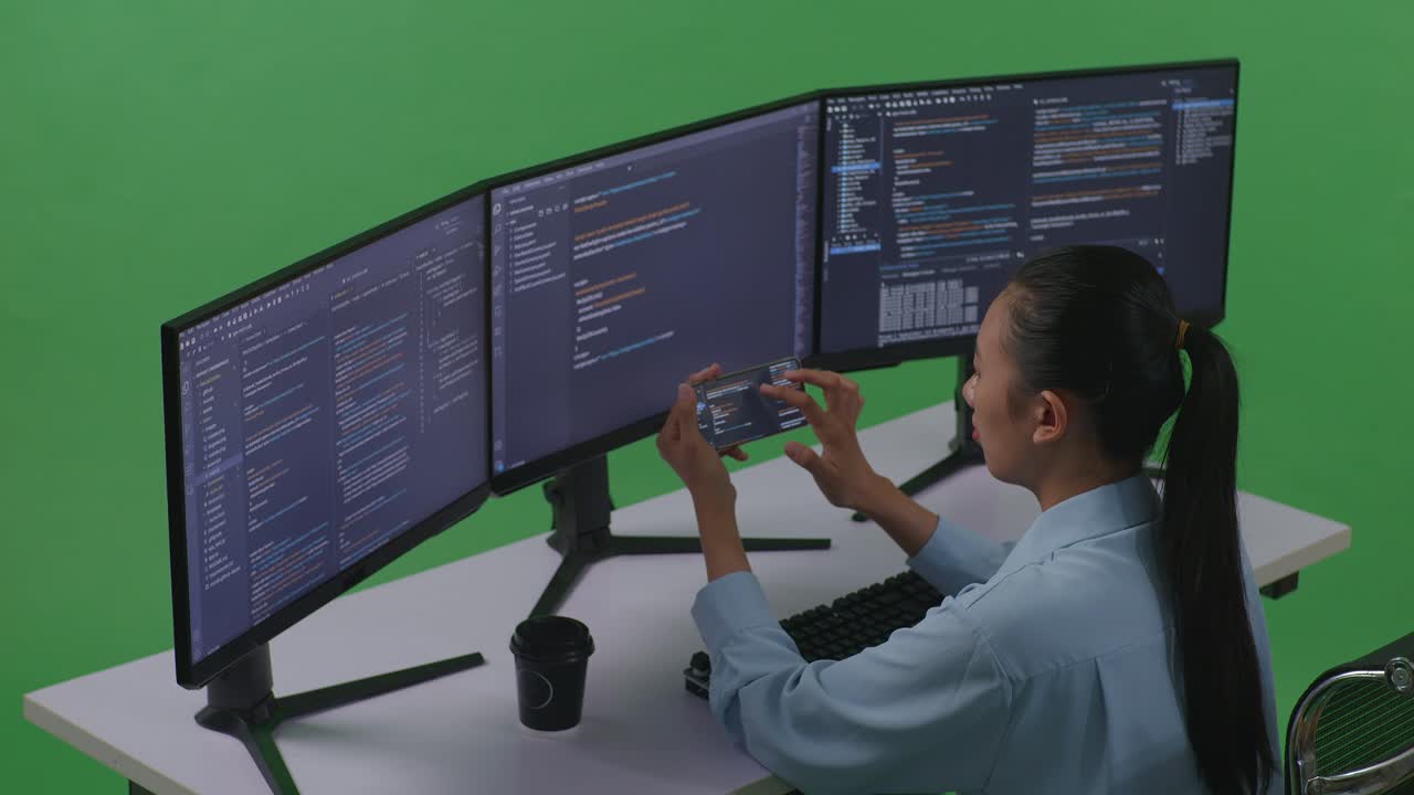 vista lateral de una desarrolladora asiática trabajando con un teléfono móvil mientras escribe código con múltiples pantallas de computadora en un estudio de pantalla verde