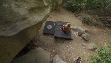 female rock climber sitting below large boulder stretching before climbing