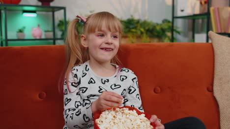 Funny-little-child-girl-watching-comedy-video-film-on-tv,-eating-popcorn-on-comfortable-sofa-at-home