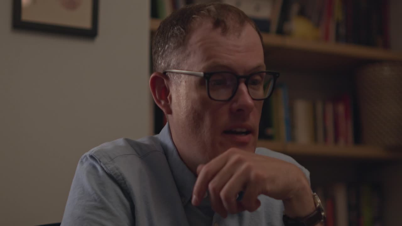 A close up of a man in glasses at work talking and laughing on a virtual meeting. Office library in the background