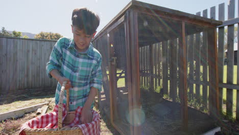 Portrait-of-happy-biracial-boy-holding-basket-with-eggs-next-to-henhouse,-slow-motion,-copy-space