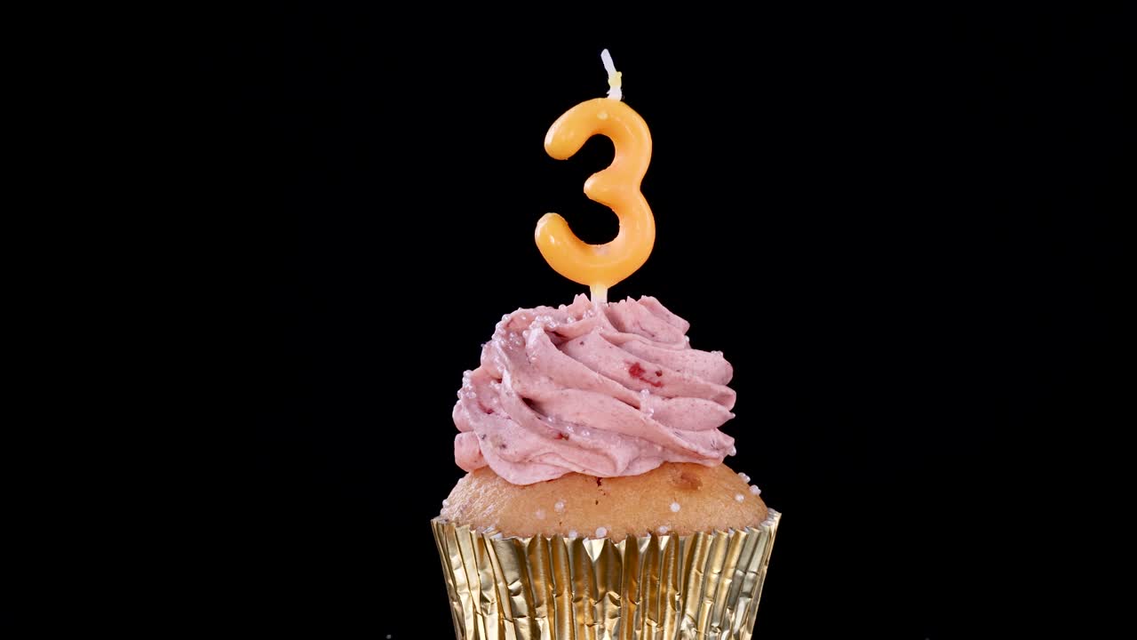 A hand inserts a yellow number three birthday candle into a strawberry frosted cupcake with gold wrapper, set against a black background under studio lighting