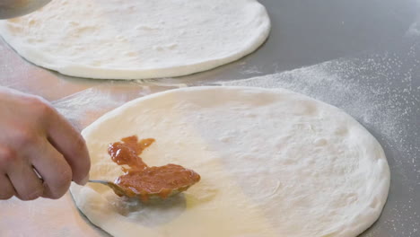 Close-Up-Of-An-Unrecognizable-Chef-Spreading-Tomato-Sauce-With-Spoon-On-The-Pizza-Raw-Dough-At-Restaurant