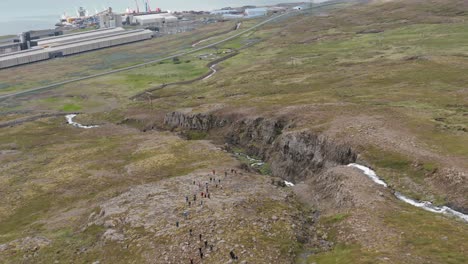 Antena-De-Grupo-De-Senderismo-Subiendo-La-Montaña-Con-La-Zona-Industrial-De-Alcoa-Fjarðaál-En-Segundo-Plano.