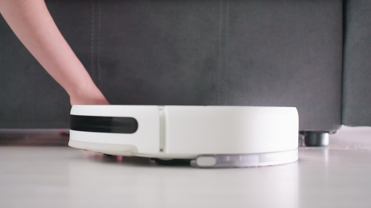 Close up of human hand stopping robot vacuum from moving forward on floor, symbolizing interaction between person and smart cleaning device