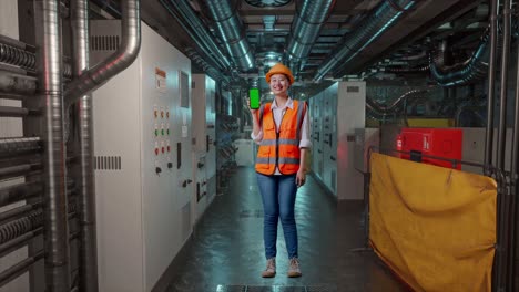 full body of asian female engineer with safety helmet smiling and showing green screen smartphone to the camera in engine control room, work of electrical generators