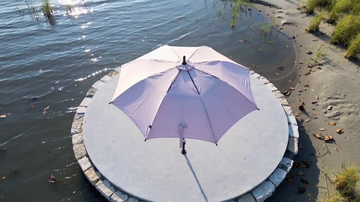 Telecamera drone che si sposta rivelando un ombrellone color lavanda su una piattaforma rotonda vicino alla riva, mostrando increspature sull'acqua.