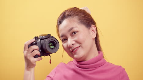 고립된 노란색 배경-1에 대해 디지털 카메라를 사용하여 분홍색 옷을 입은 행복한 활기 넘치는 아시아 여성