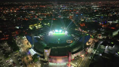 Filmische-Drohnenaufnahme-Von-Blinkenden-Lichtern-Im-Stadion,-Während-Eine-Musikband-Nachts-Auf-Der-Bühne-Steht-–-Eine-Menge-Feiernder-Fans-In-Santo-Domingo-–-Beleuchtetes-Stadtbild-Im-Hintergrund,-Dominikanische-Republik