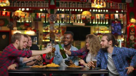 In-a-multi-ethnic-company-of-friends-a-girl-offers-a-toast-and-friends-banging-glasses-and-bottles-of-beer-while-sitting-in-a-bar.-A-fun-evening-in-the-company-of-friends.