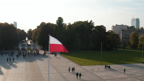 Orbit-shot-around-national-flag-of-Poland-waving-on-pole-on-Pilsudski-Square.-People-walking-across-tiled-square-of-monument-in-rest-of-Saxon-Palace.-Warsaw,-Poland