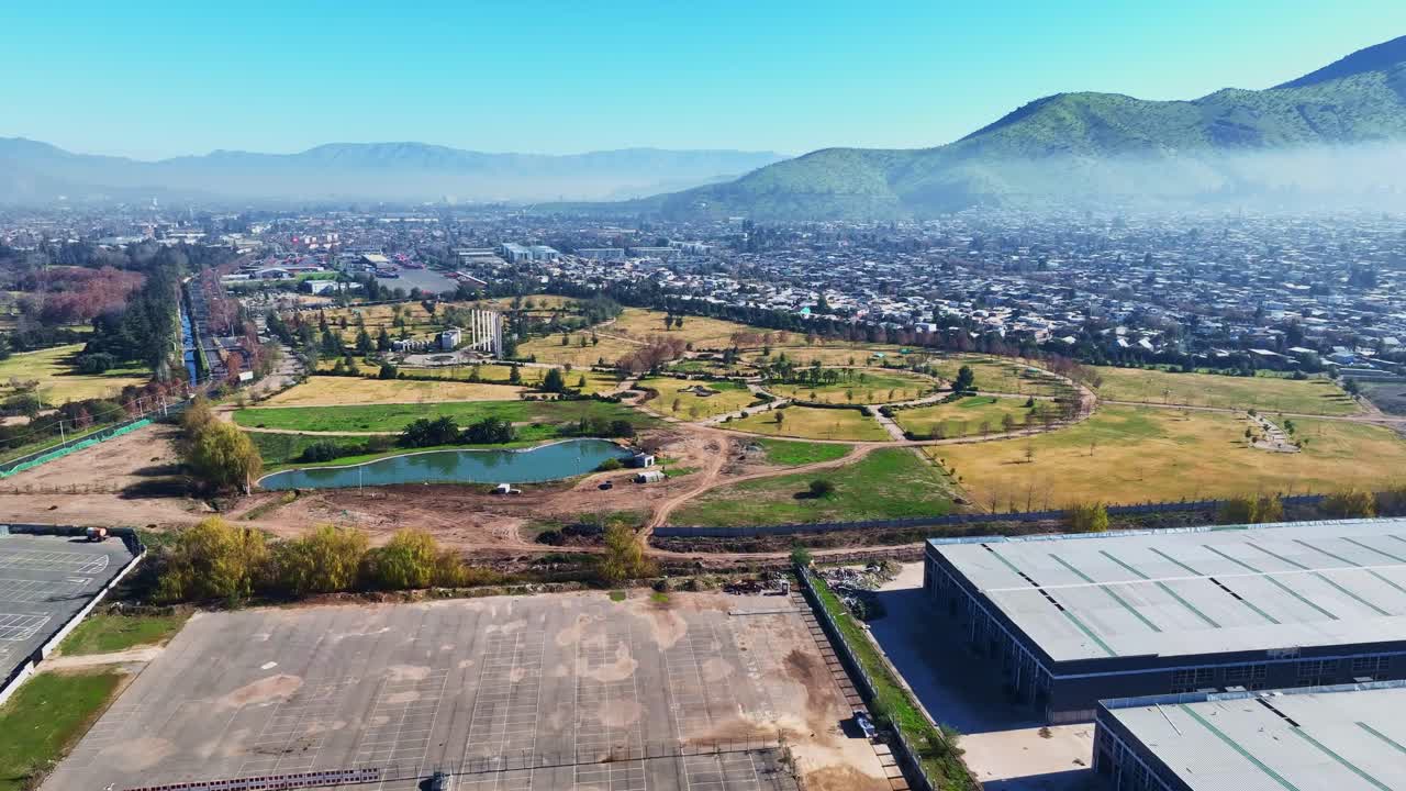 Forward drone aerial of urban park with lake and pathways, Santiago, Chile with mountains, residential neighborhoods and hazy atmosphere