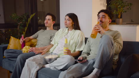 side view of three friends watching interesting movie on television sitting on couch, eating popcorn and chips and drinking soda