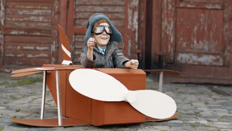 niño feliz con pelo rojo en sombrero y gafas sentado al aire libre en un modelo de avión de juguete de madera y soñando con ser aviador