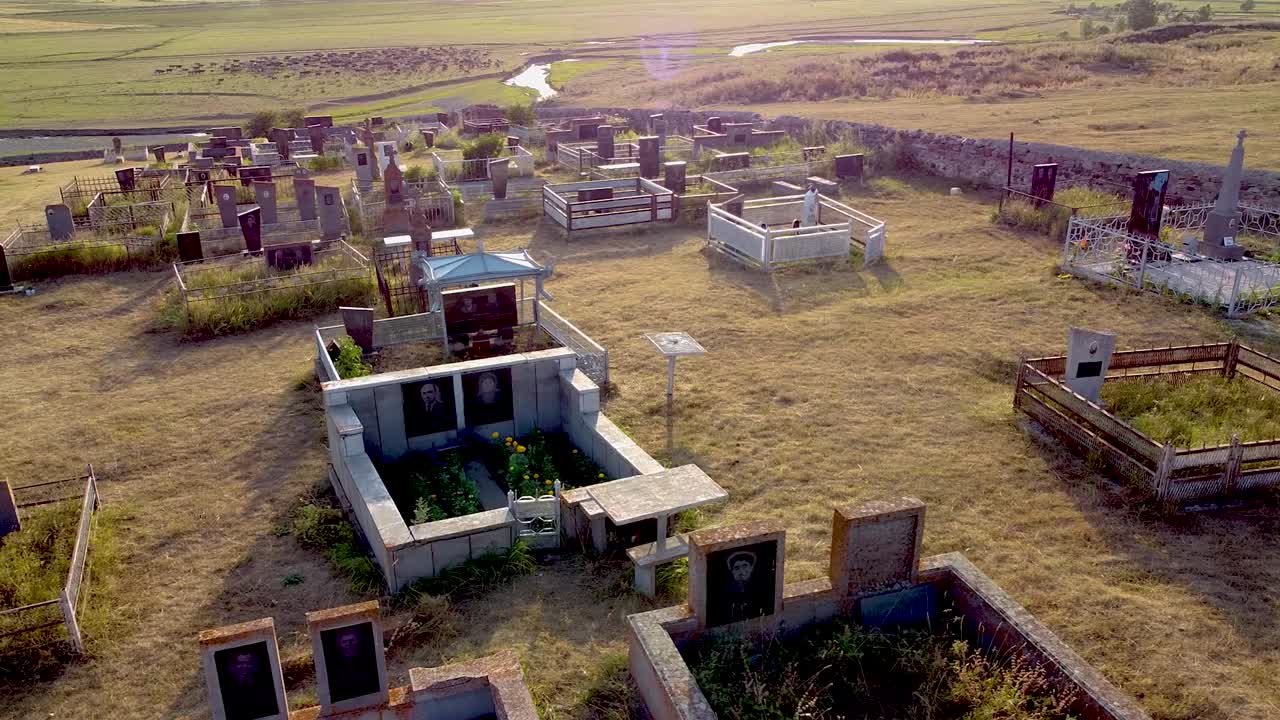 An aerial view reveals an abandoned cemetery featuring distinctive graves. The setting sun casts a warm glow over the historic site surrounded by vast fields and serene streams
