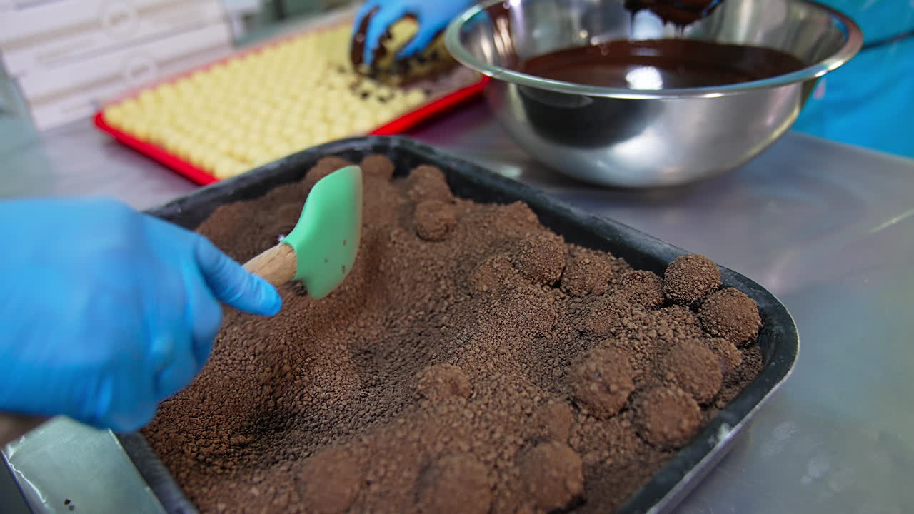 Covering chocolate candies with cocoa crumbs. Gloved hand mixes sweets with spatula. Handmade chocolate desserts close up.