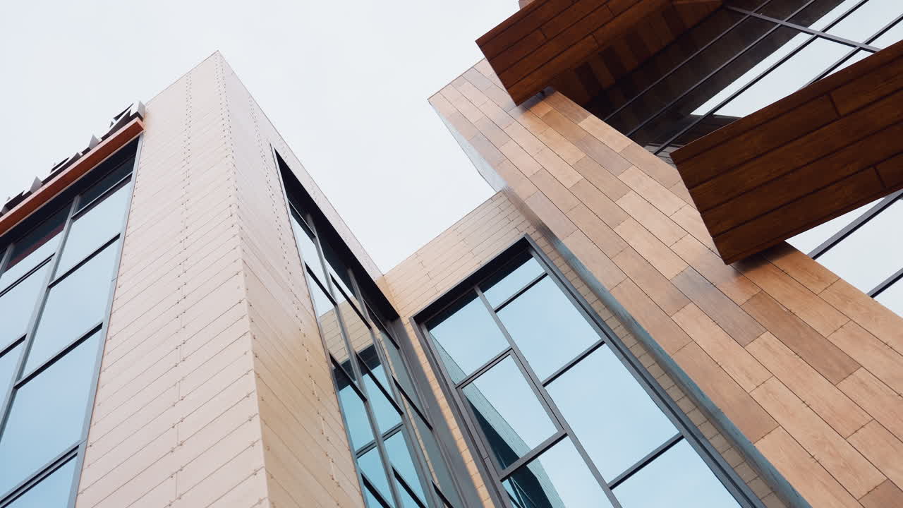 Upward angle view of modern fitness building featuring large glass windows and wooden panel facade under bright daylight sky, showcasing sleek architecture and vertical lines in urban setting