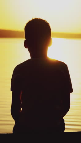 Little boy sitting on wooden dock at sunset. Child is watching the sunset against the background of a river. Dreams Come True. Vertical video