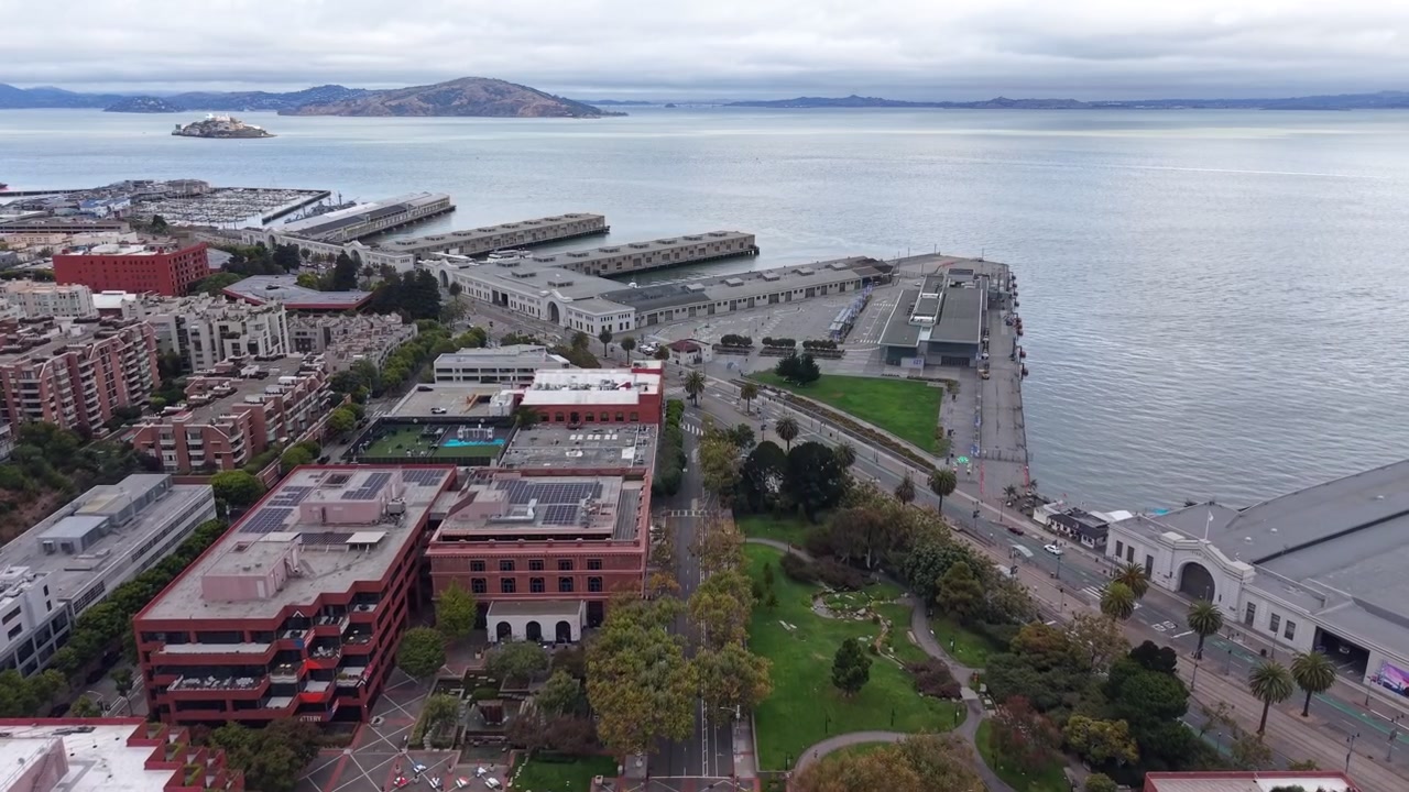 Veduta aerea nuvolosa che rivela l'iconica Isola di Alcatraz, la Baia di San Francisco e l'architettura urbana del lungomare dell'Embarcadero, inclusi i suoi moli storici e gli edifici circostanti della città