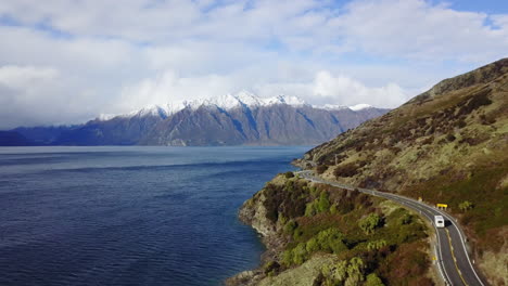 Aerial-Tracking-shot-of-RV-in-New-Zealand-Driving-on-a-coastal-road-with-snowy-mountains-in-background-in-4K