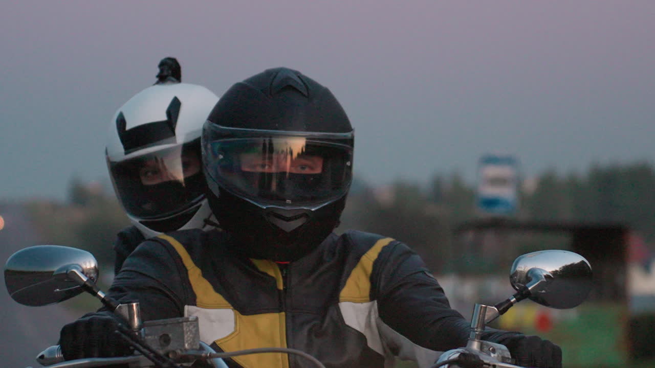 Close up of couple wearing protective helmets riding motorcycle uphill on countryside road during twilight, hands on handlebar, headlight glowing, relaxed mood, open air, freedom, long road ahead