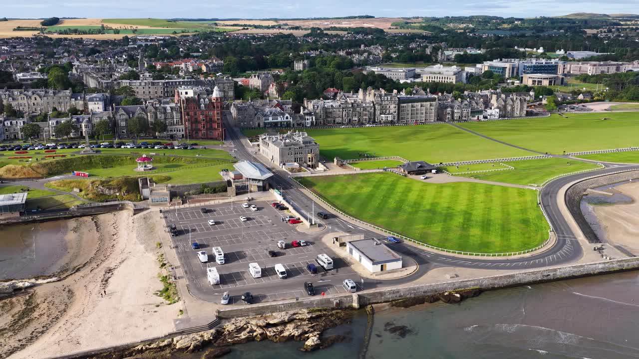 Drone pans above coastal golf course, historic clubhouse, parking lot, and town under bright daylight