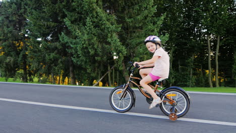 funny girl five years old riding a bicycle with extra wheels smiling