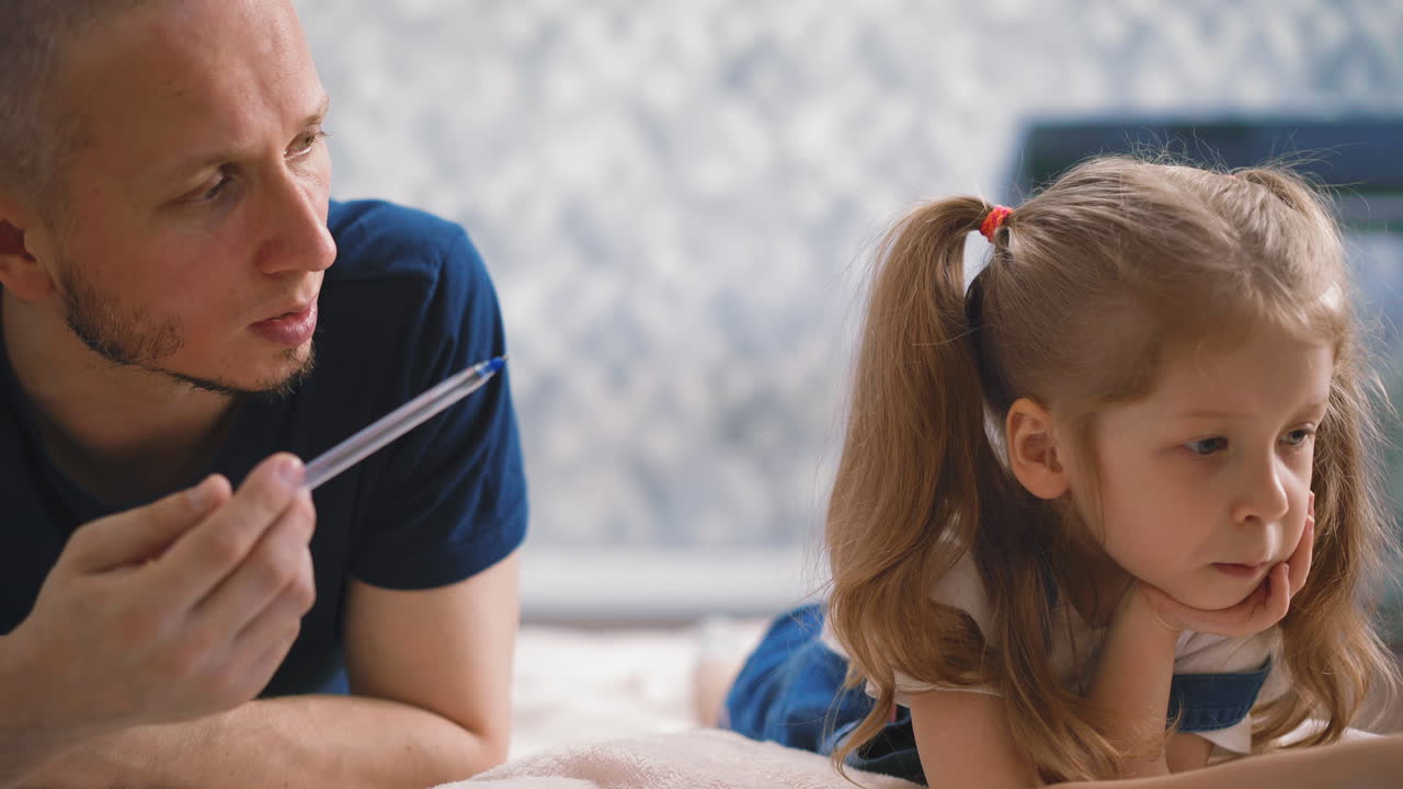 아빠는 바닥에 누워 있는 금발 소녀에게 숙제를 설명합니다.