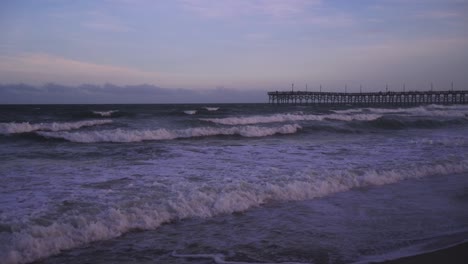 Olas-En-Una-Playa-En-Cámara-Lenta-Con-Un-Muelle-De-Madera-Al-Fondo