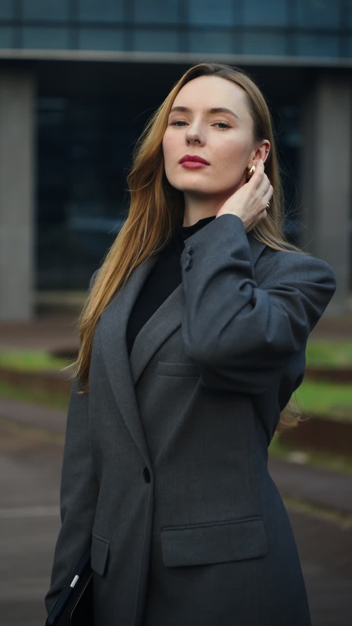 A confident Caucasian businesswoman looks towards the camera and then turns into profile, adjusting her hair behind her ear as the camera slowly dollies in on =office building terrace