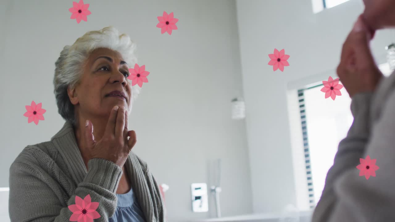 animación de flores rosadas sobre feliz mujer mayor biracial poniendo maquillaje en el espejo en casa