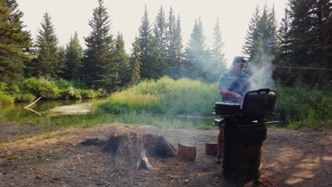 wide angle view of man grilling steaks while camping by small pond