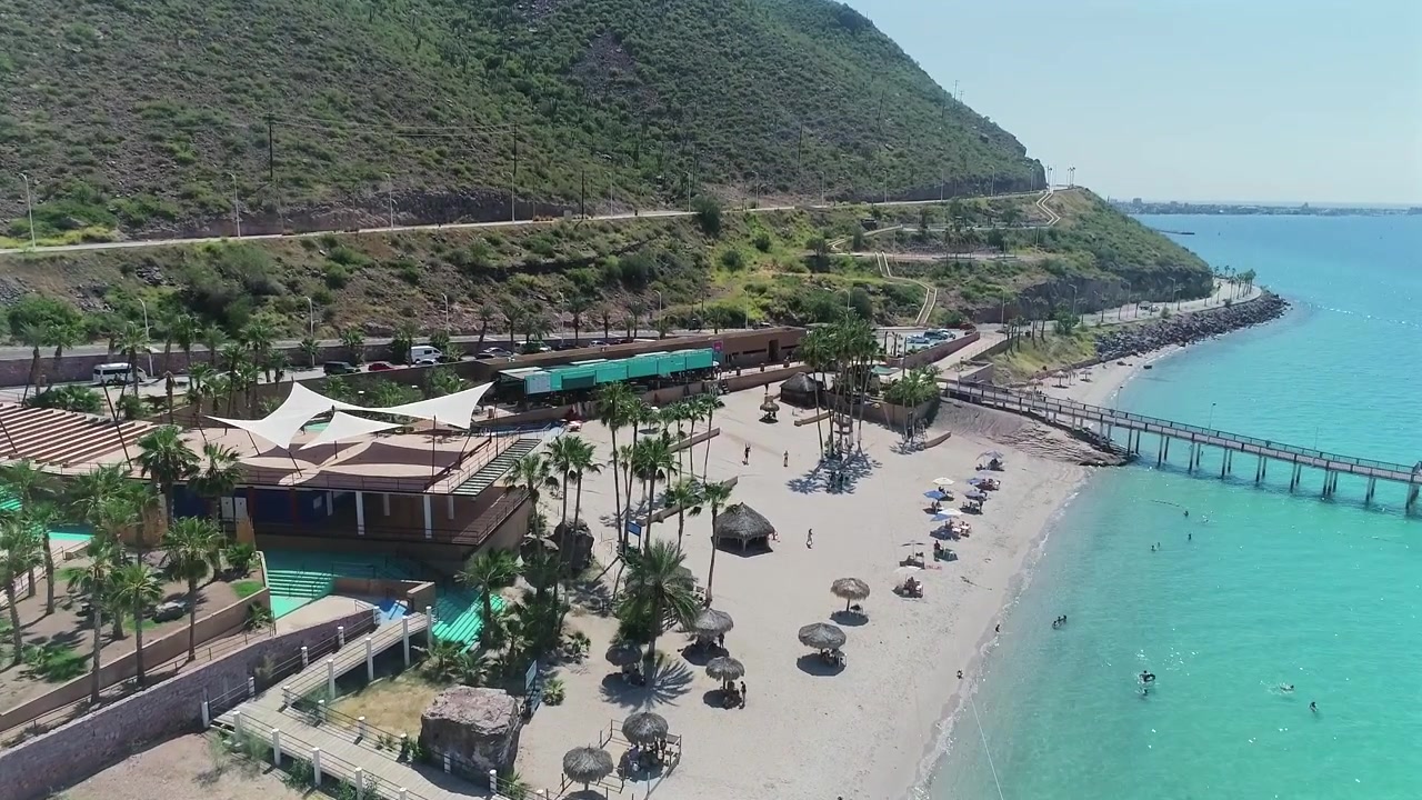 Cảnh quay drone 4K từ trên không tiến vào từ phía đại dương hướng ra bãi biển, tiết lộ một lối đi bộ trên mặt nước, một cầu trượt nước và các khu vực nghỉ ngơi