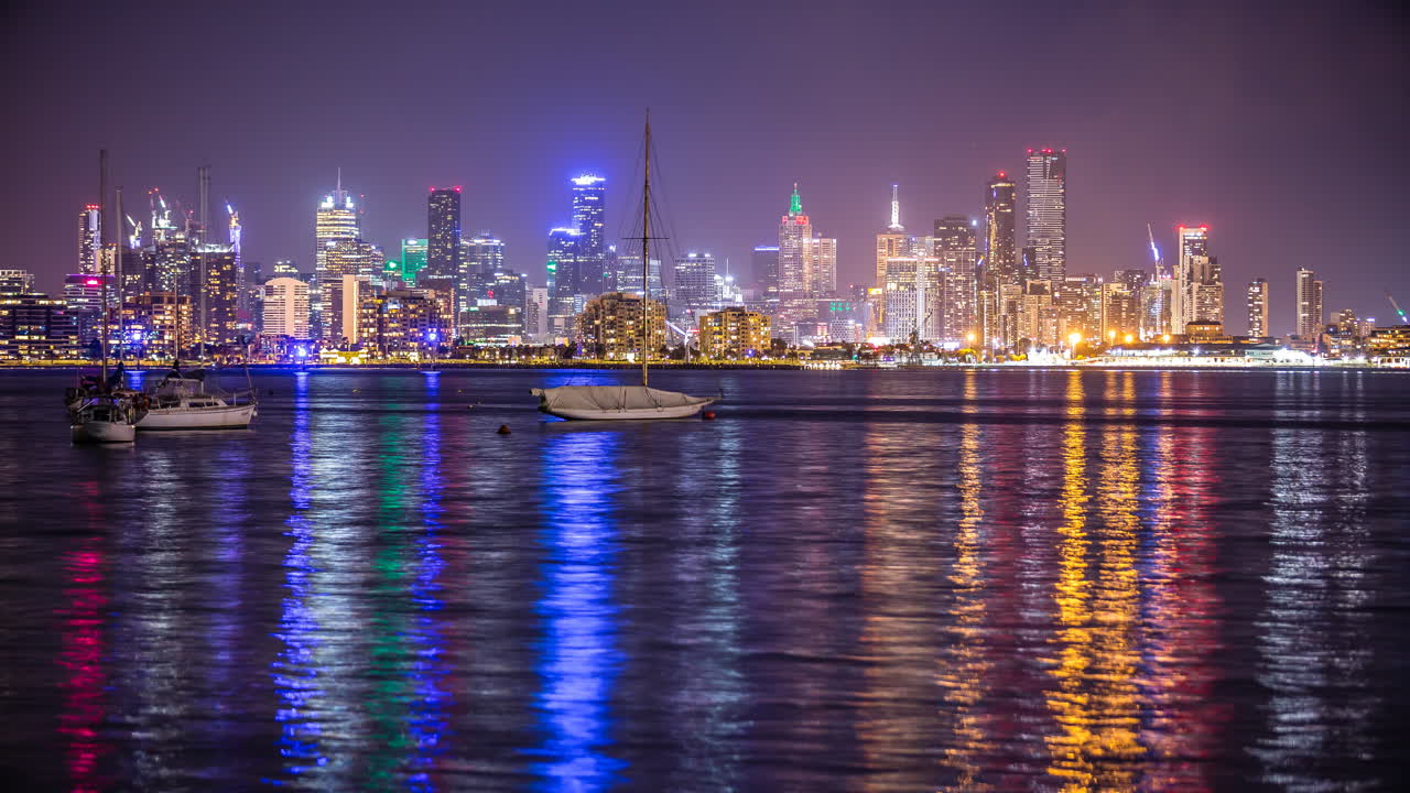 Timelapse notturno di Melbourne, Australia, sull'acqua con riflessi colorati che mostra i grattacieli della città da Williamstown.