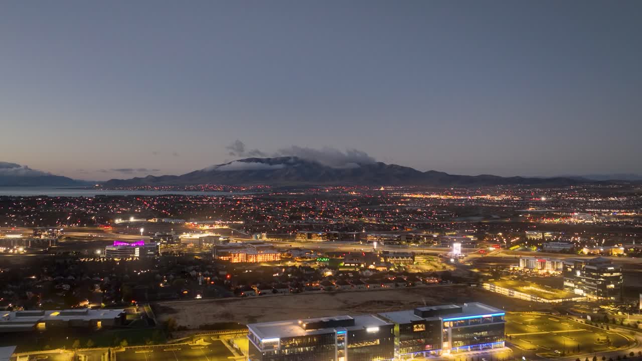 Aerial footage of Utah Valley and Lake viewed from Silicon Slopes in Lehi at dawn. The video captures the tranquil landscape and glowing city lights against a mountain backdrop