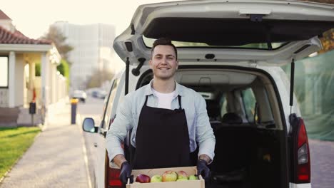 friendly smiling man with braces holding wooden box with apples at street outdoors, portrait of happy delivery man in apron and