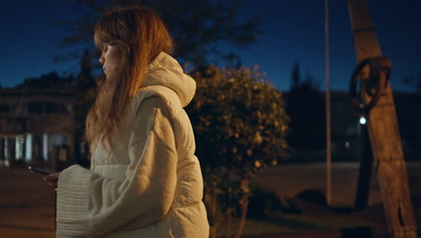 young woman using smartphone in illuminated urban park at night closeup.